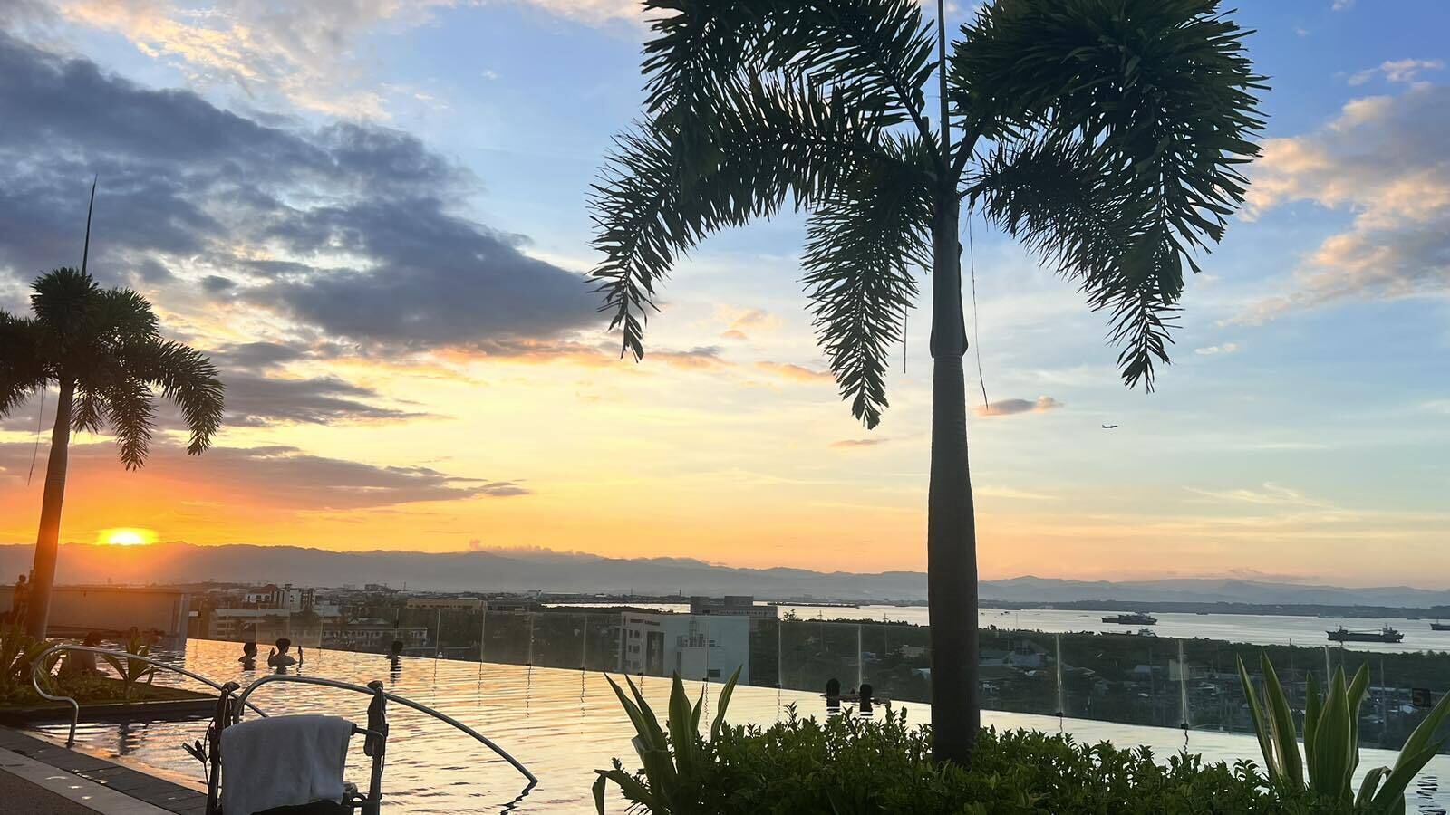 A sunset view from a balcony overlooking a body of water with ships, palm trees, and distant mountains.