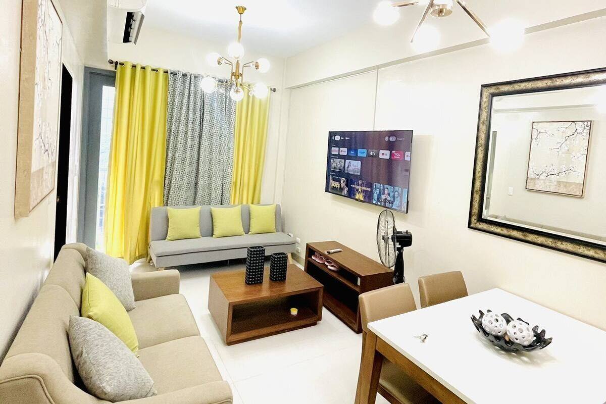 A modern living room with a sofa, coffee table, dining table, and a flat-screen TV mounted on the wall.