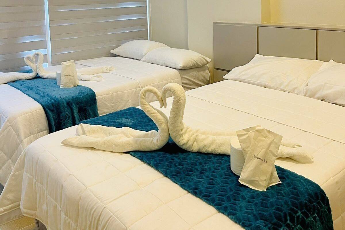 A hotel room with two beds, each with a white duvet and a teal throw blanket. There are two white towels folded on each bed, and a white bag labeled "Essential Kit" on the left bed.