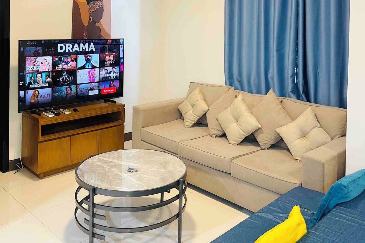 A living room with a television displaying a drama channel, a beige sofa, a blue curtain, and a small round table.