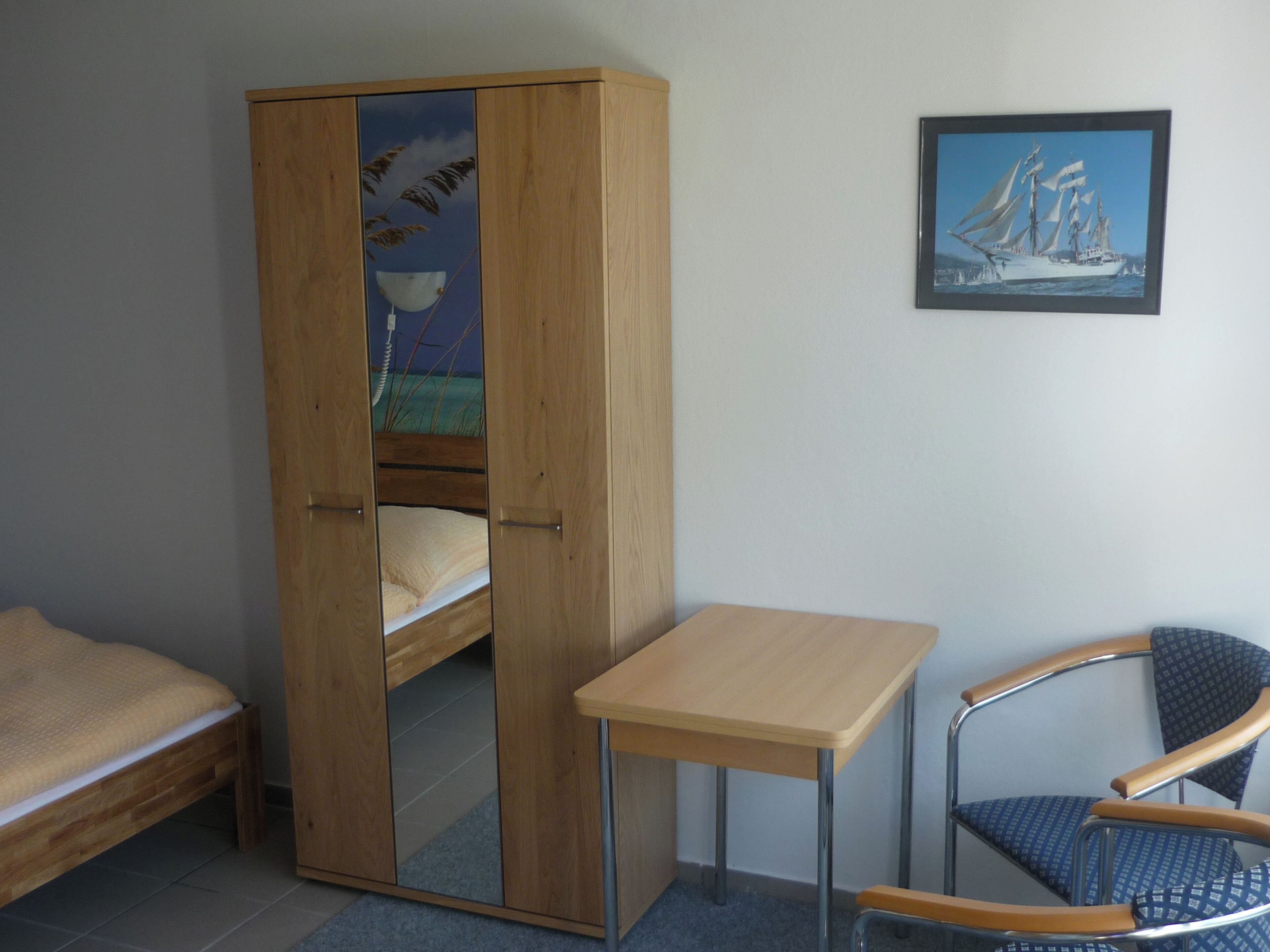 A bedroom with a wooden wardrobe, a bed, a small table, and a chair.