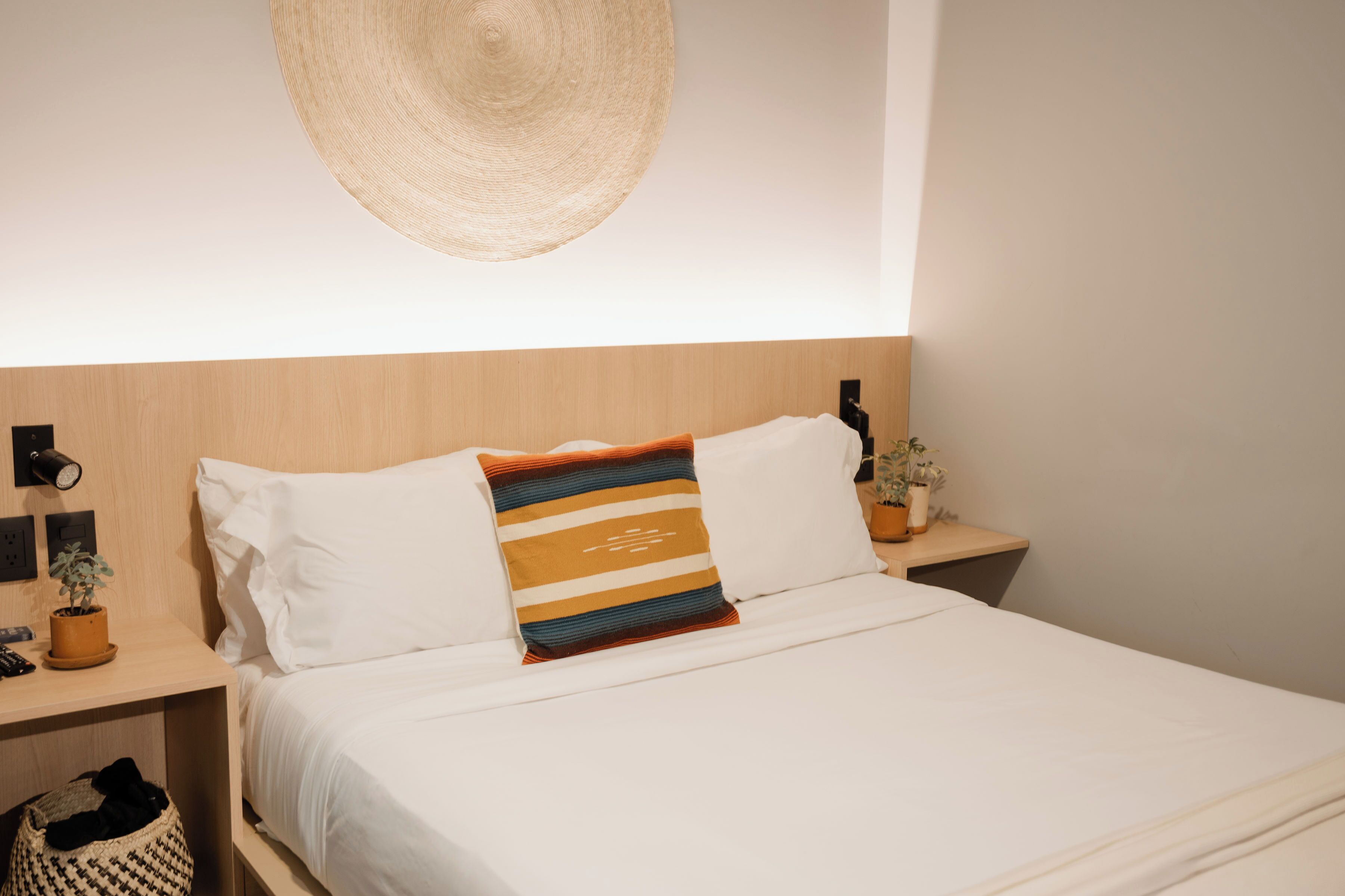 A neatly made bed with white linens and striped pillows, a woven wall hanging above, and a small plant on a bedside table.