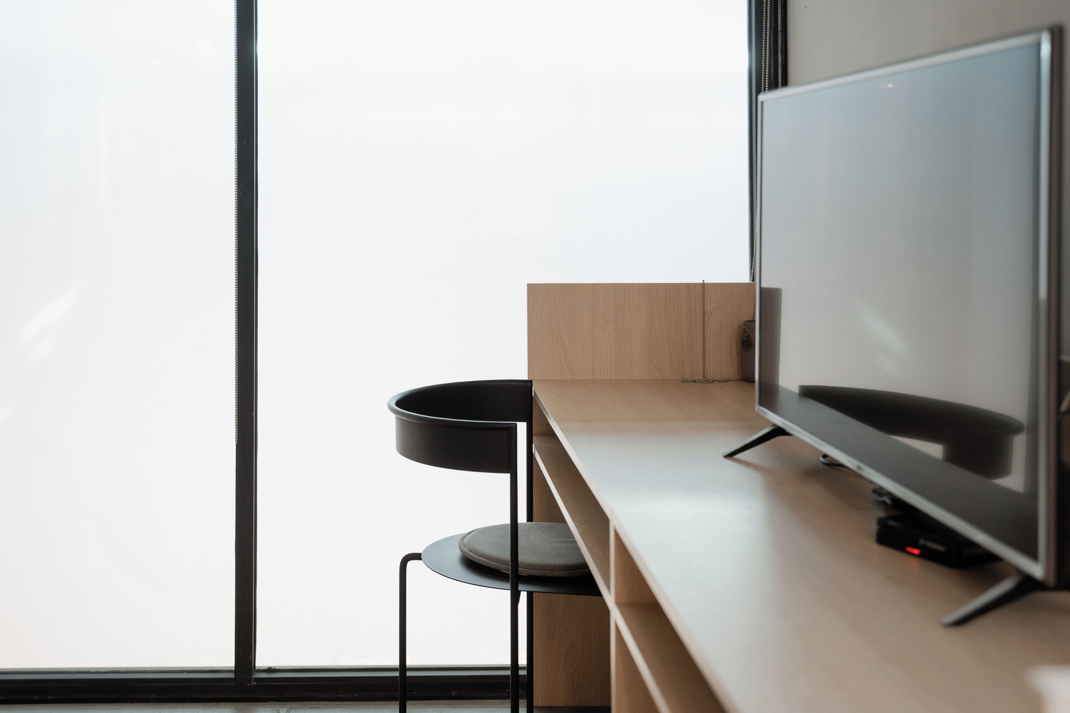 Una habitación moderna con un ventanal grande, un televisor de pantalla plana sobre un atril de madera y una silla negra.