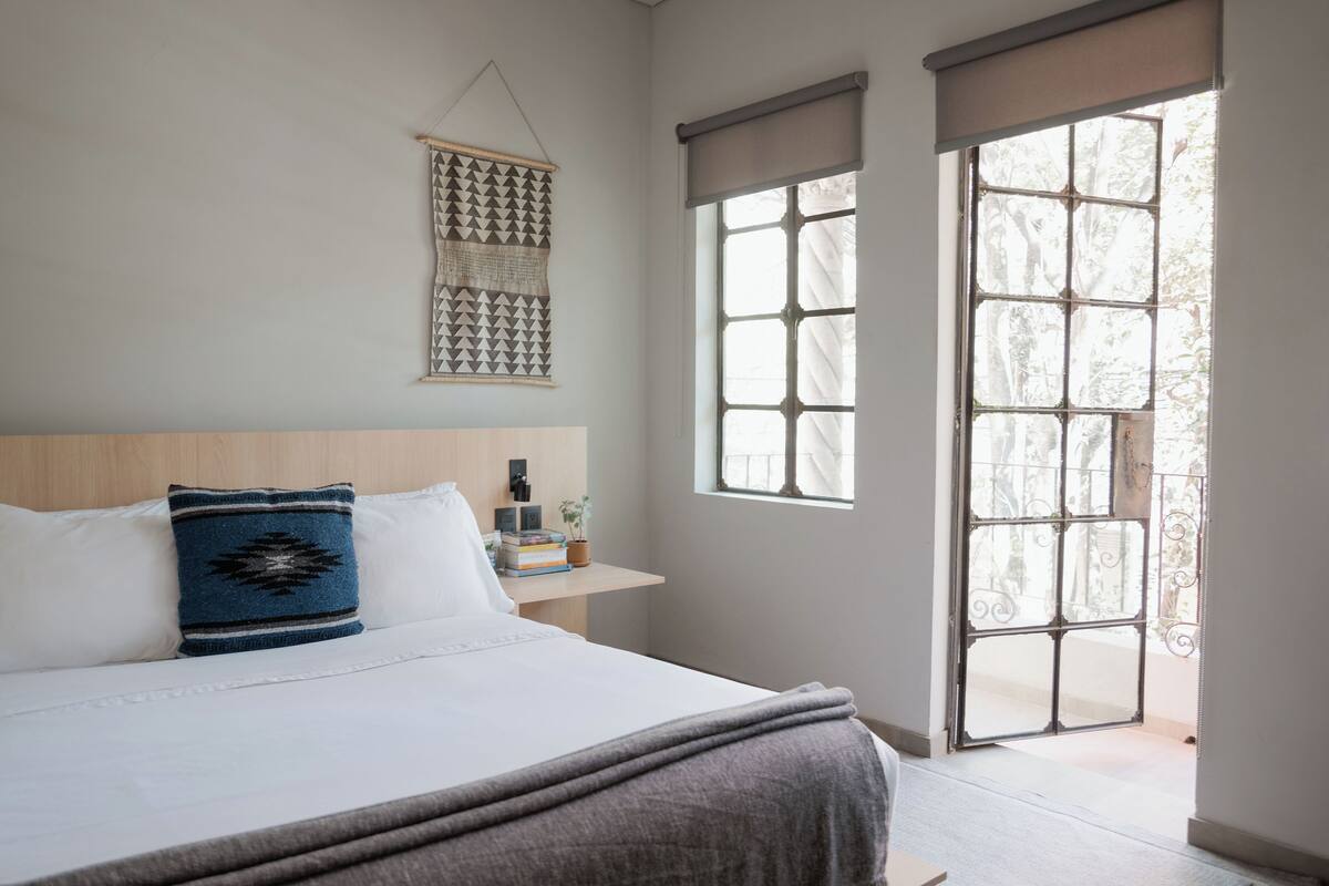 A bedroom with a bed, pillows, a bedside table with books and a plant, a wall hanging, and a large window with a view of trees.