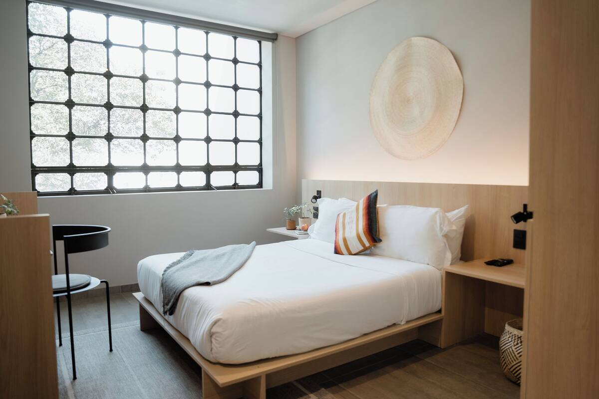 A modern bedroom with a large window, a bedside table, a bedside lamp, and a wall-mounted artwork.