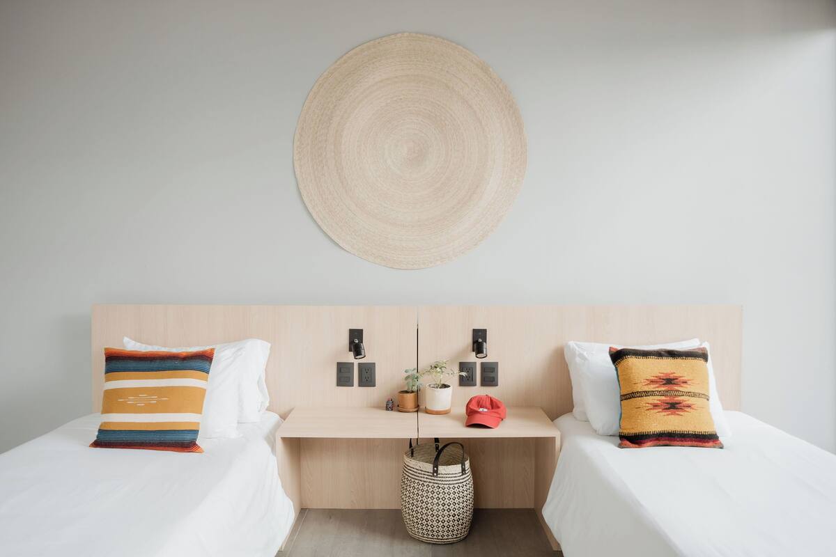 A hotel room with two beds, a wooden desk, a woven wall hanging, and a small potted plant.
