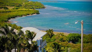 Vista costera con un edificio de techo curvo, rodeado de exuberante vegetación y un mar azul cristalino.