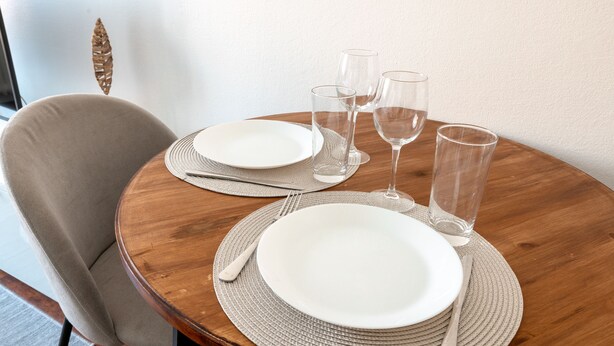 Una mesa de madera redonda con platos blancos, cubiertos y vasos transparentes, y un camino de mesa tejido debajo.