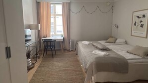 Un dormitorio con cama, escritorio y silla. Hay una ventana con cortinas, un radiador y una lámpara fijada a la pared.