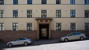 Un edificio de varios pisos con una entrada central, dos coches aparcados y una calle empedrada.