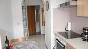 A kitchen with a stove, oven, and coffee maker. There is a table with a bottle and a red object. A clock is on the wall. - holiday apartment Serlesblick (Fulpmes)
