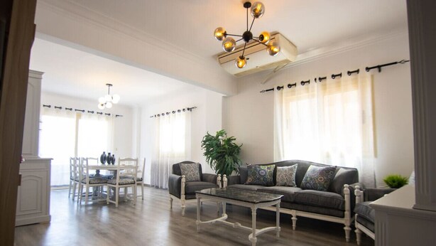 A spacious living room with a dark-colored sofa, a coffee table, and a dining area with a white table and chairs. - Lovely 3BR Apartment in Heart of Mohandseen (Giza Governorate)