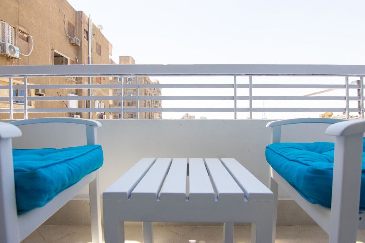 Un balcone con due sedie bianche e un tavolino, con vista su un edificio con diverse finestre e condizionatori.