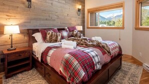 A bedroom with a wooden wall, a large window with a mountain view, a bed with plaid bedding, and two wall-mounted lamps.
