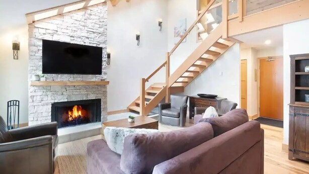 A modern living room with a stone fireplace, a flat-screen TV, a staircase, and a cozy seating area.
