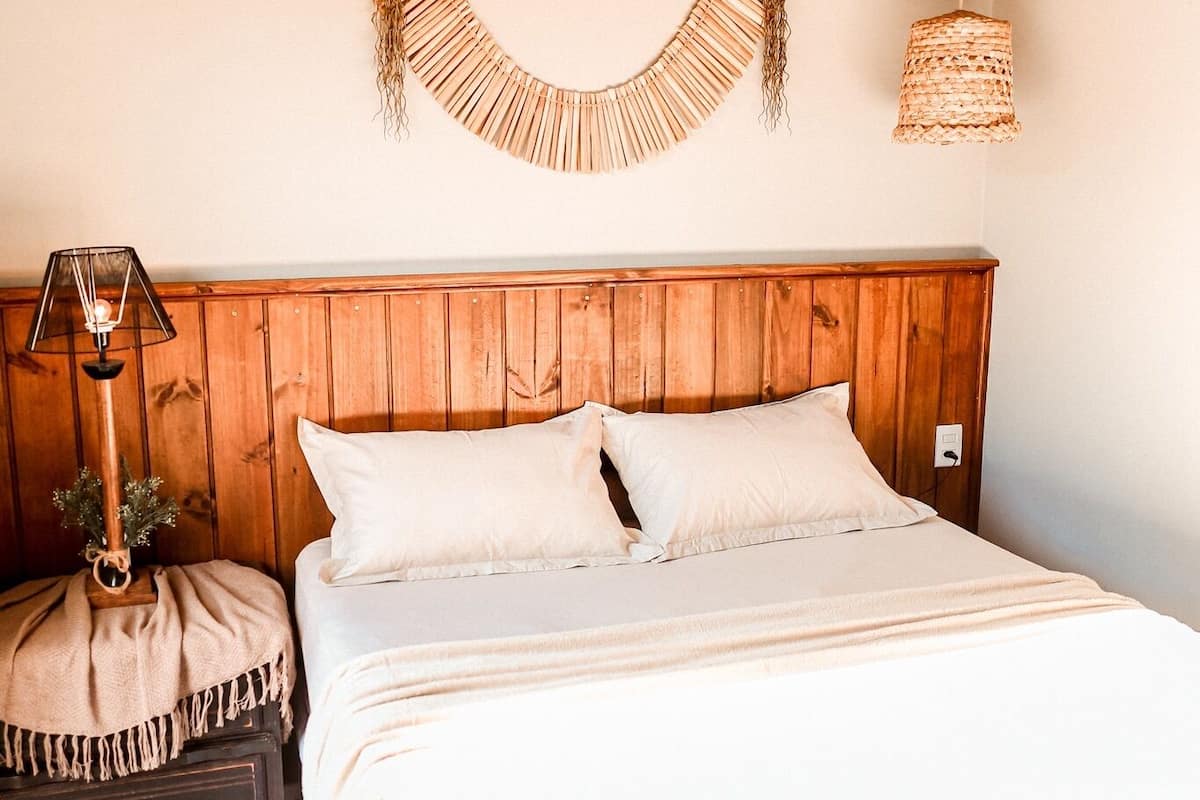 Une chambre à coucher avec une tête de lit en bois, un lit recouvert de draps blancs, une lampe de chevet et une décoration murale tissée.