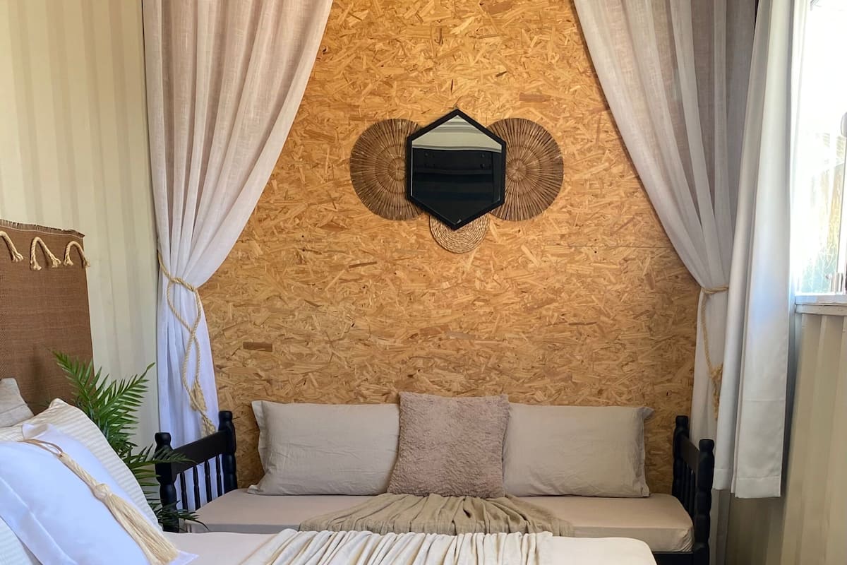 Une chambre avec un mur en bois, un lit recouvert d’une couverture beige et des rideaux blancs.