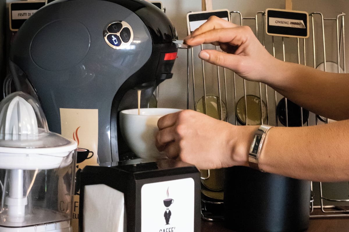A coffee machine with a label reading "CAFFÈ CASTELLANO" and a hand holding a cup. There are oranges in a wooden bowl on the counter.