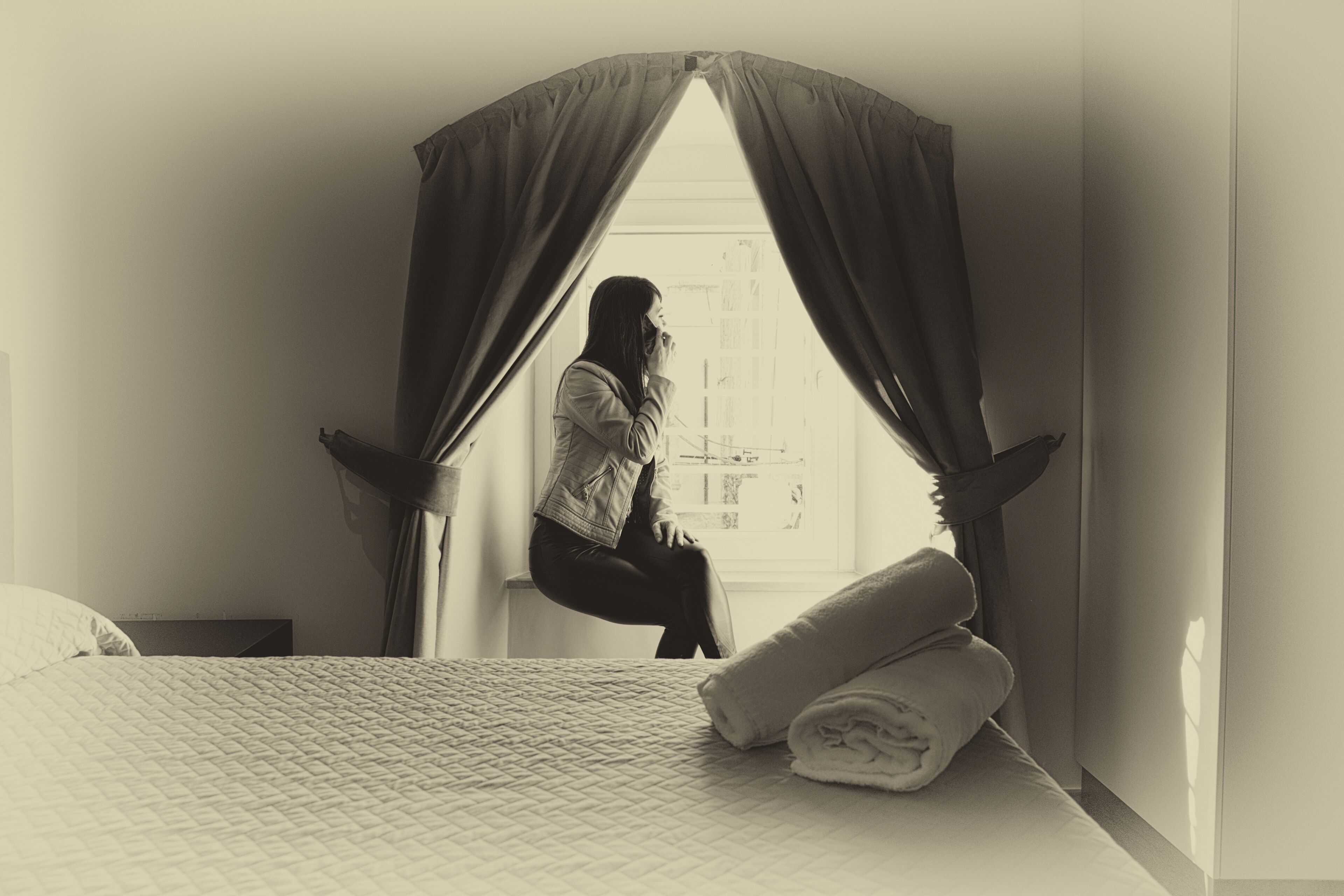 A bedroom with a bed, a window with curtains, and a person sitting by the window.