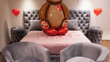 Une chambre à coucher avec une tête de lit capitonnée, un grand lit recouvert d’une couverture rose, deux fauteuils gris et un ours en peluche en guise de décoration.