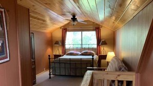 Un dormitorio con techo de madera, una cama con cabecera, dos lámparas de noche, una ventana con cortinas y un pasamanos de madera al frente.