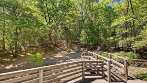 Un puente de madera que conduce a un área de picnic con mesas y sillas, rodeado de árboles y una pequeña masa de agua.