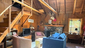 L’intérieur d’un chalet accueillant, avec un escalier en bois, une cheminée en pierre, un téléviseur et un canapé bleu.
