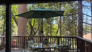 A covered outdoor deck with a round glass table and chairs, surrounded by wooden railings and large trees.