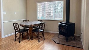 Un coin repas avec une table ronde et quatre chaises, une fenêtre avec des stores et un poêle à bois noir.