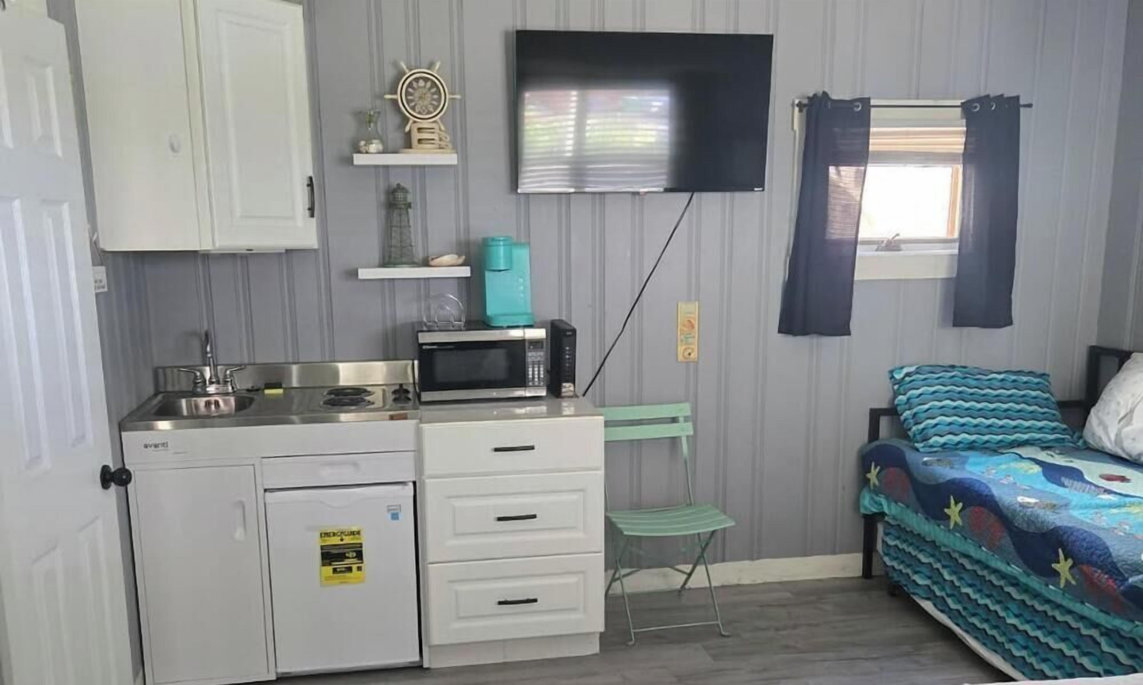 A compact room with a kitchenette, a bed with a patterned bedspread, a television mounted on the wall, and a window with curtains.