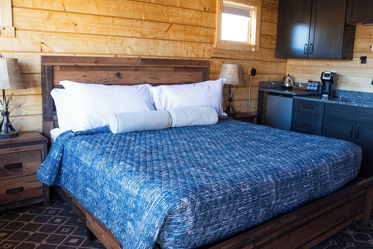 A wooden cabin bedroom with a bed, a ceiling fan, a window, and a kitchenette.