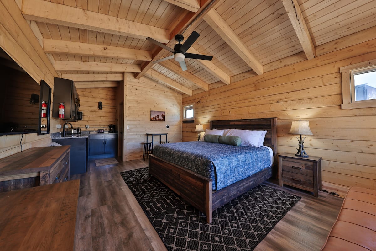 A wooden cabin interior with a bed, a desk, a kitchenette, and a ceiling fan.
