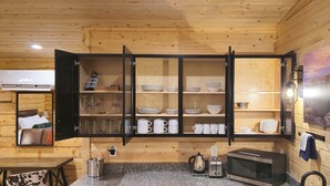 A compact kitchen with wooden walls, a built-in cabinet with dishes, a kettle, a microwave, and a small dining area. - Muddy Creek Mining Company (Hanksville)