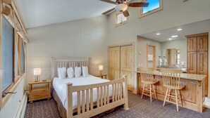 Un dormitorio con cama de madera, ventilador de techo, escritorio y dos sillas, y un baño visible a través de una puerta abierta.