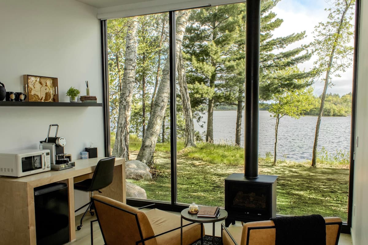 A modern room with a large window overlooking a lake, a wooden desk with a microwave, a chair, and a small table with a book and a bowl of fruit.