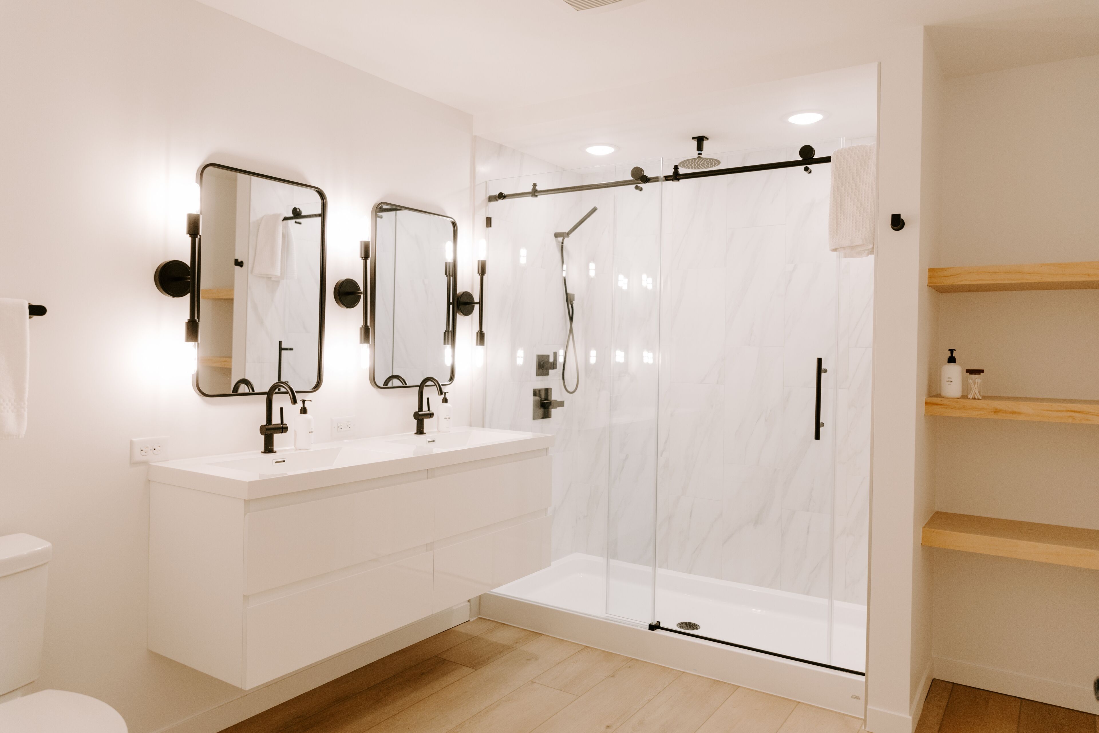 Un baño moderno con un tocador blanco, dos espejos, ducha con cerramiento de vidrio y estantes de madera.