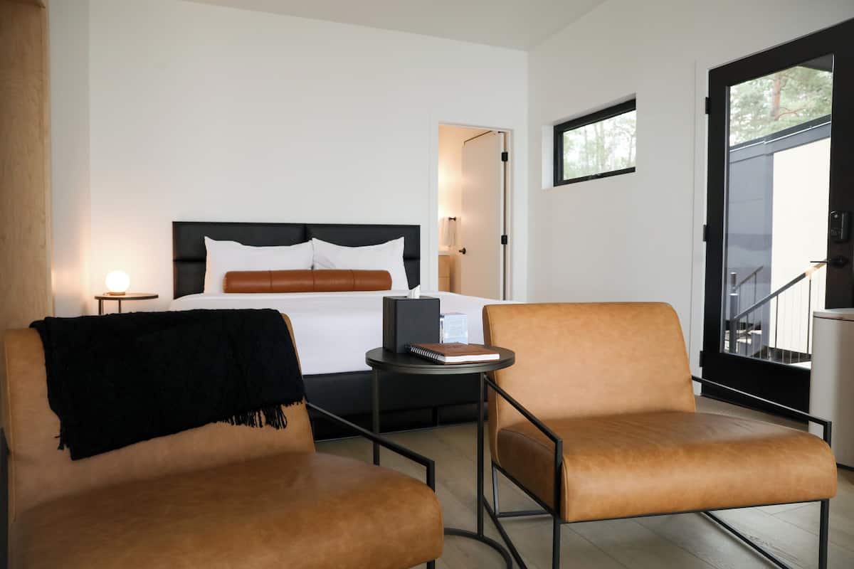 A modern hotel room with a bed, two armchairs, a small table, and a door leading to a balcony.
