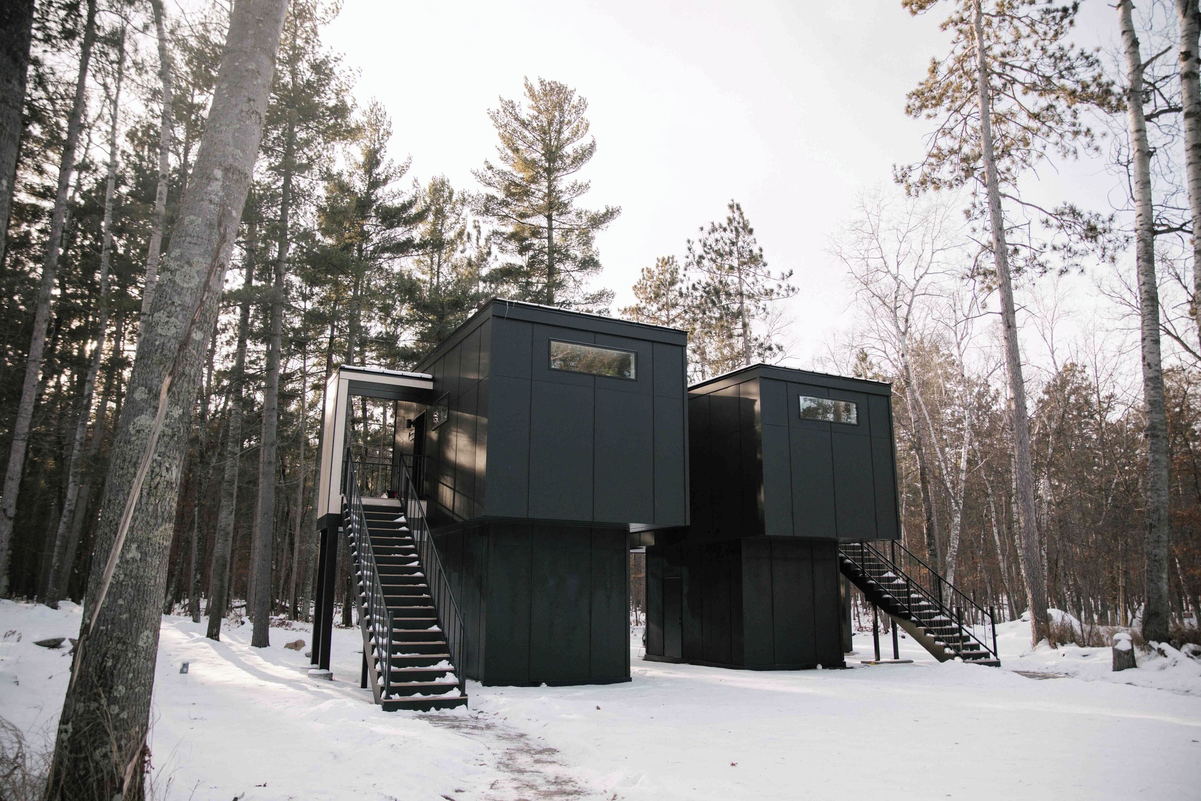 A modern cabin with a staircase leading to a deck, surrounded by a snowy forest.