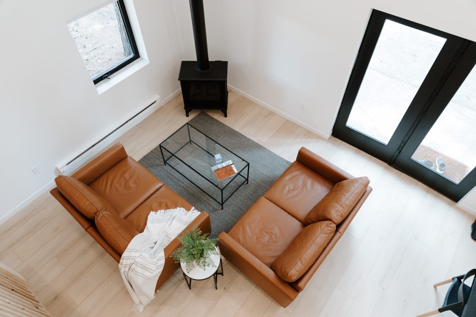 Een moderne woonkamer met een bruine leren bank, een glazen salontafel en een klein bijzettafeltje met een plant. Er is een zwarte haard zichtbaar in de hoek.