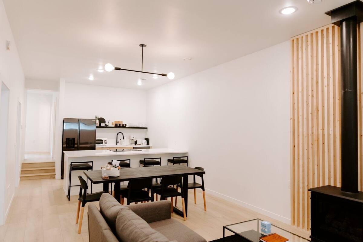 A modern living room with a sofa, dining table, and chairs. A kitchen with a refrigerator and microwave is visible in the background.