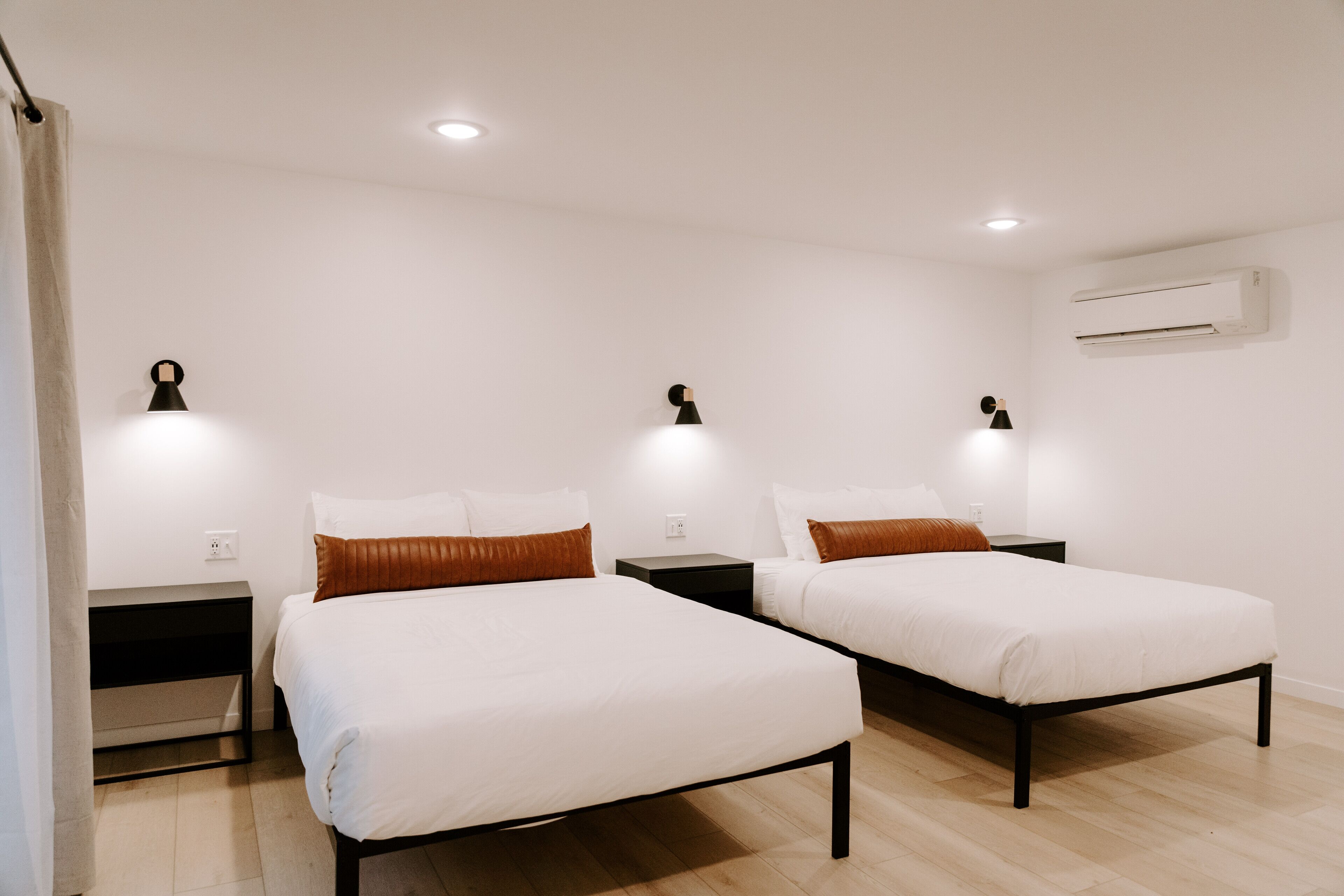 A hotel room with two single beds, a nightstand with a lamp, and an air conditioning unit on the wall.