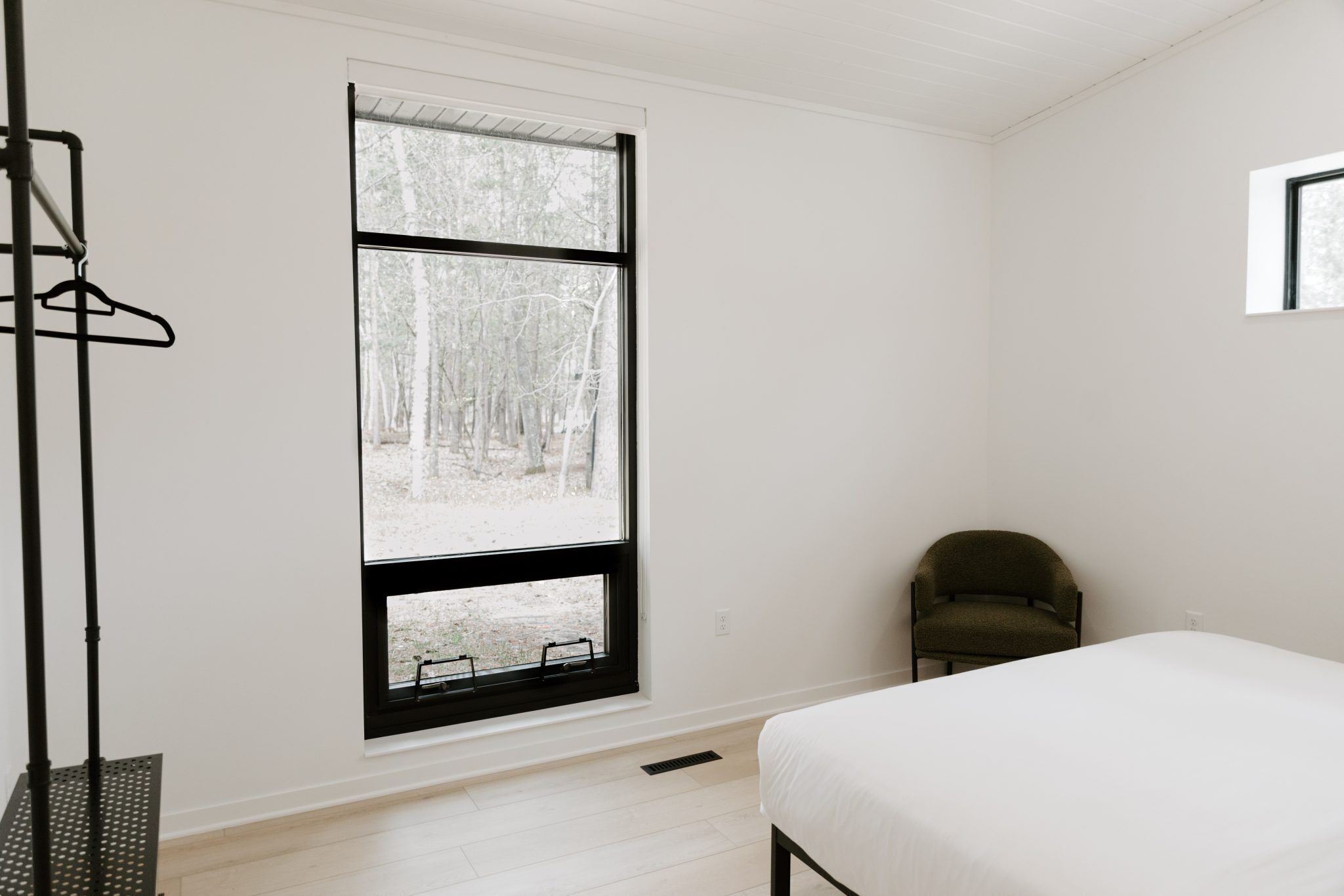 Un dormitorio con un ventanal grande, una cama, un sillón y un perchero.