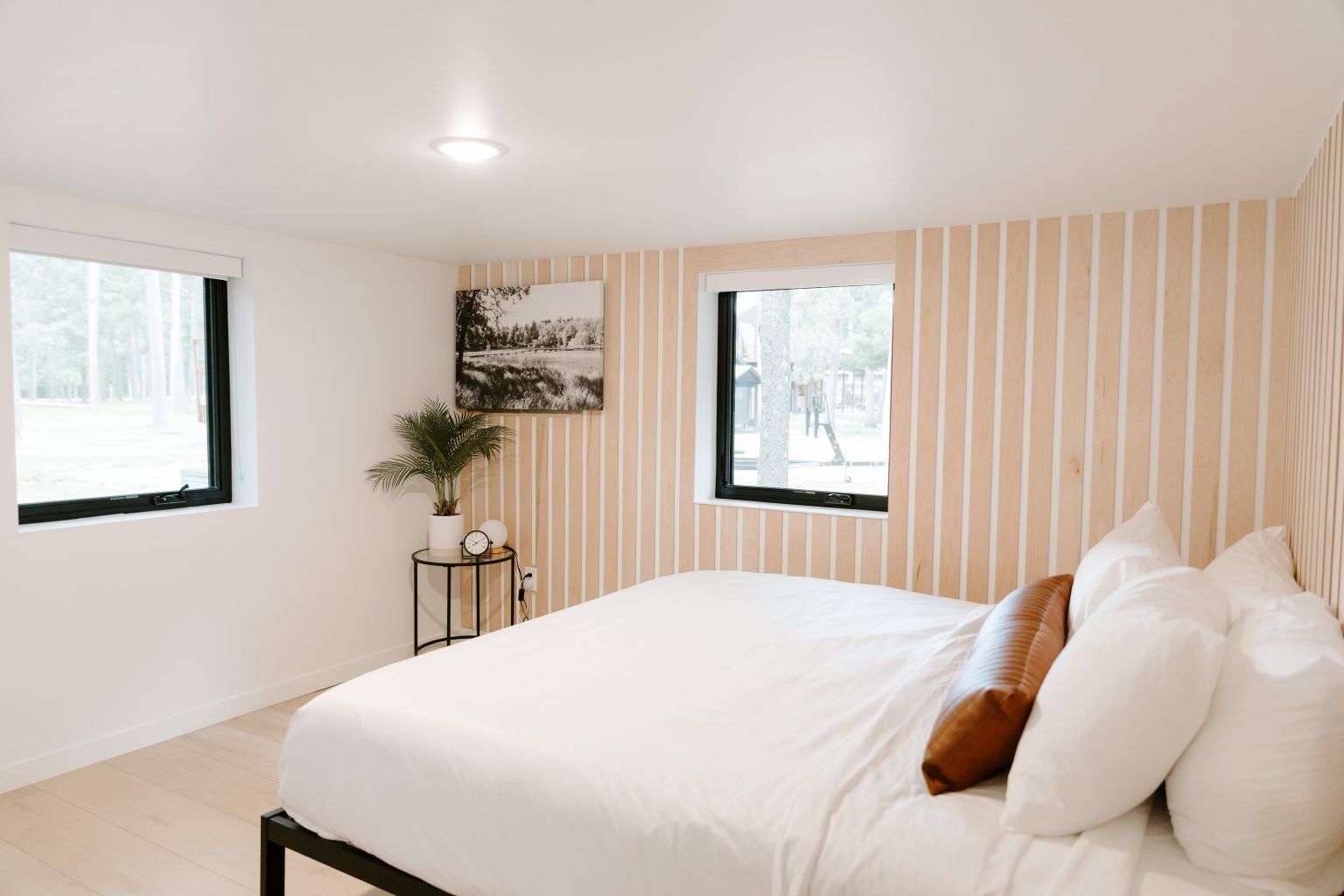 A bedroom with a bed, two windows, a wall-mounted picture, a small table with a plant and clock, and a ceiling light.