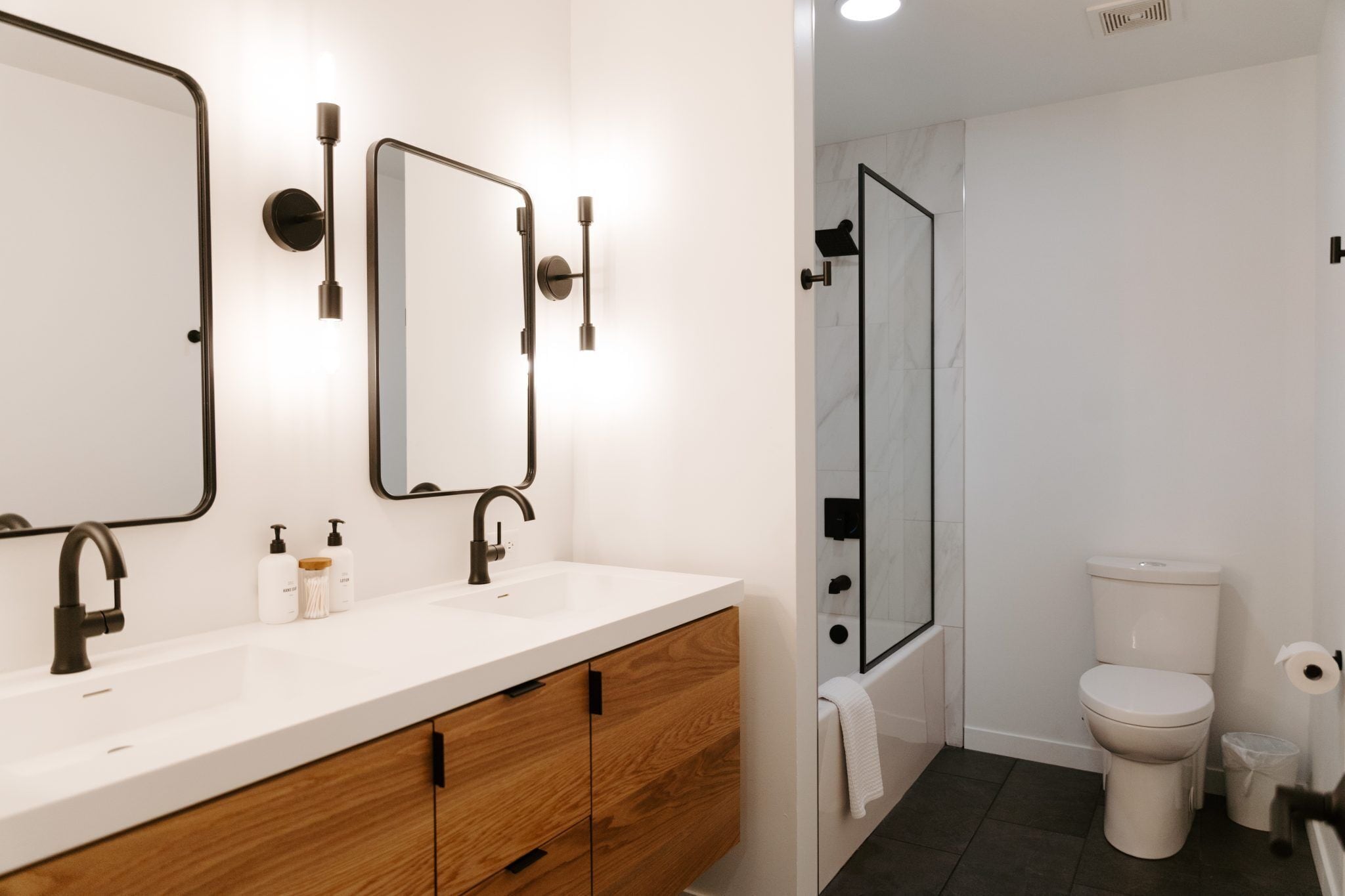 A modern bathroom with a white sink, wooden cabinets, two mirrors, a toilet, and a shower area.