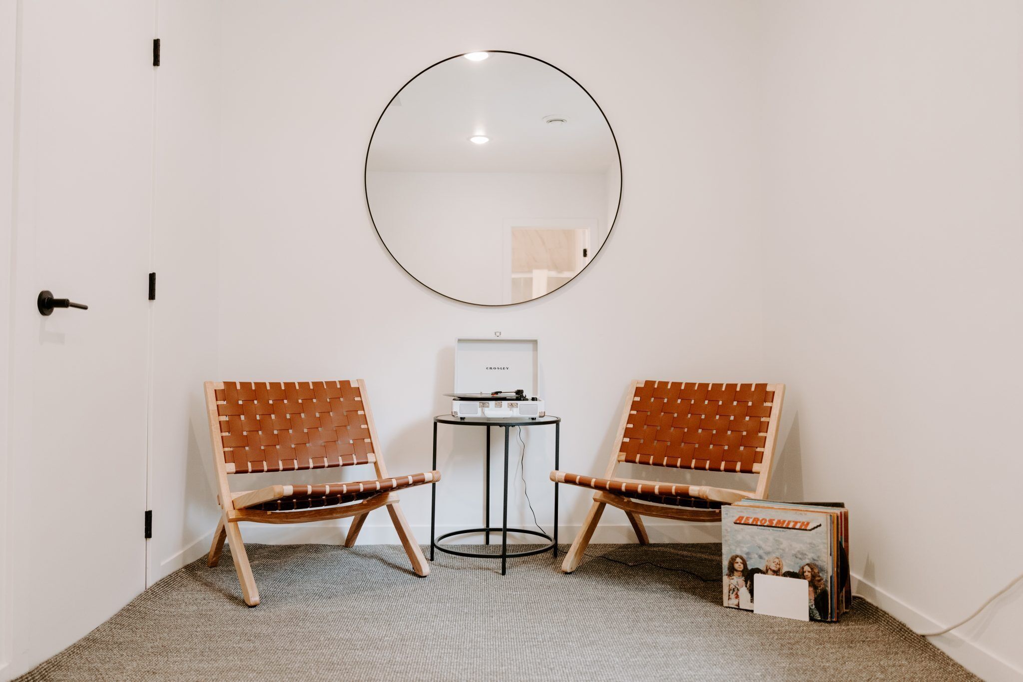 A room with two wooden chairs with brown leather seats, a round mirror on the wall, a small table with a turntable, and a framed album on the floor.