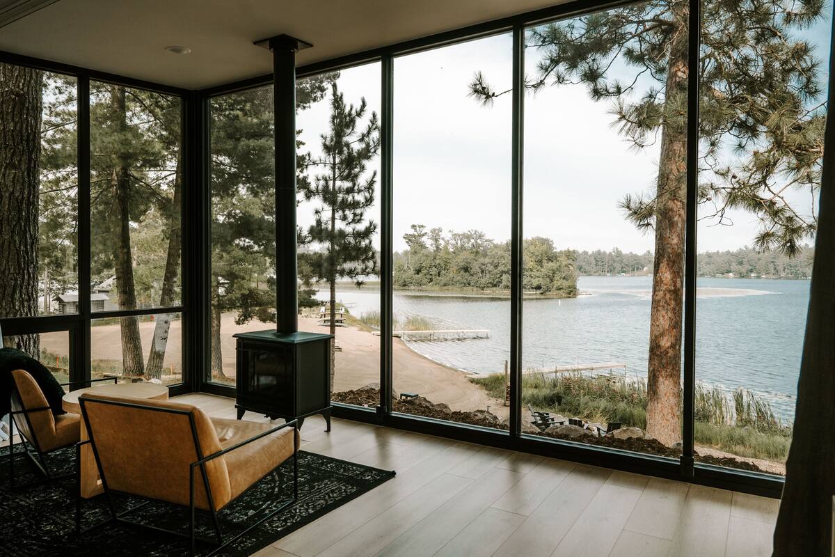 A modern room with large windows overlooking a lake, a cozy seating area, and a fireplace.