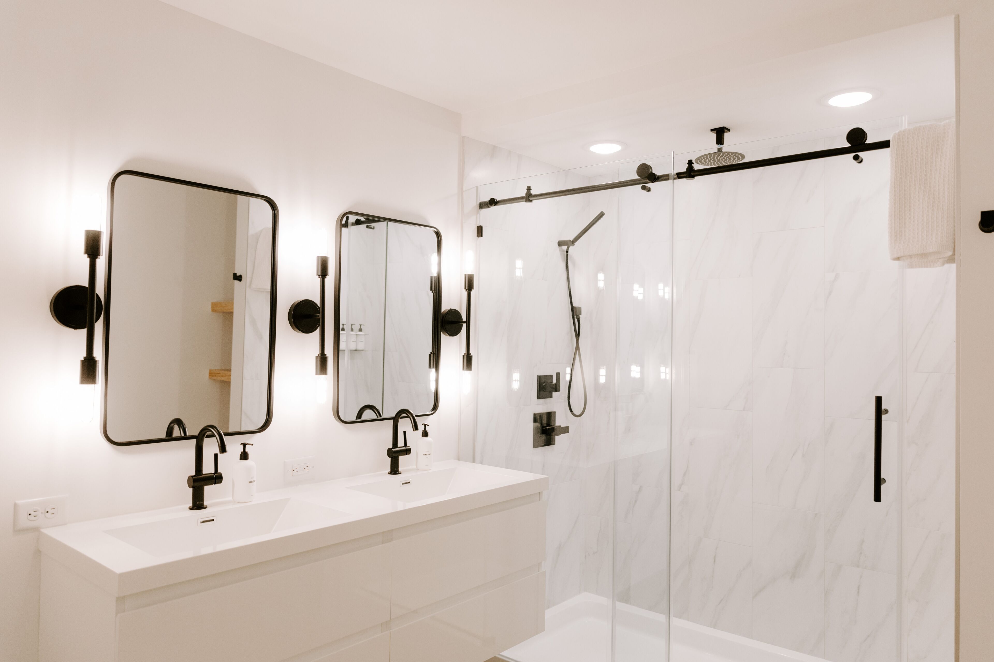 A modern bathroom with a white marble shower enclosure, a double sink vanity with black fixtures, and two wall-mounted mirrors.