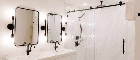 A modern bathroom with a white marble shower enclosure, a double sink vanity with black fixtures, and two wall-mounted mirrors.