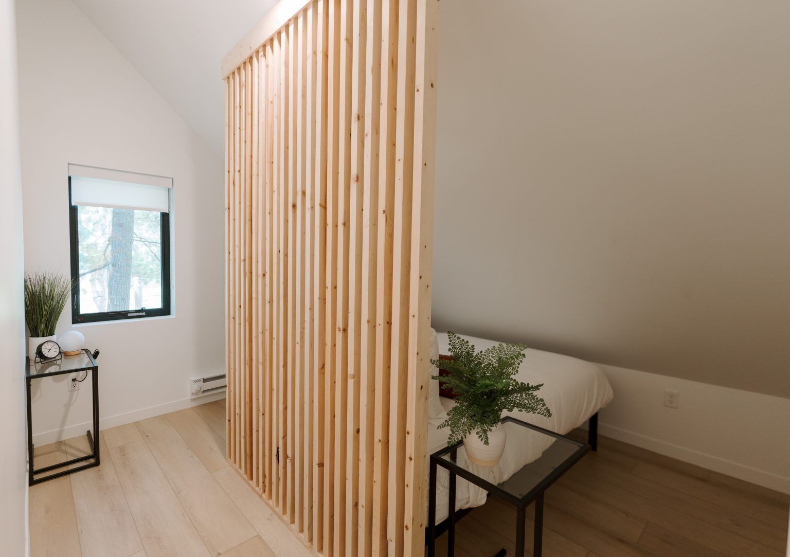 Een moderne kamer met een houten lambrisering, een raam met uitzicht op bomen, een tafeltje met een plant en een bank met een kussen.