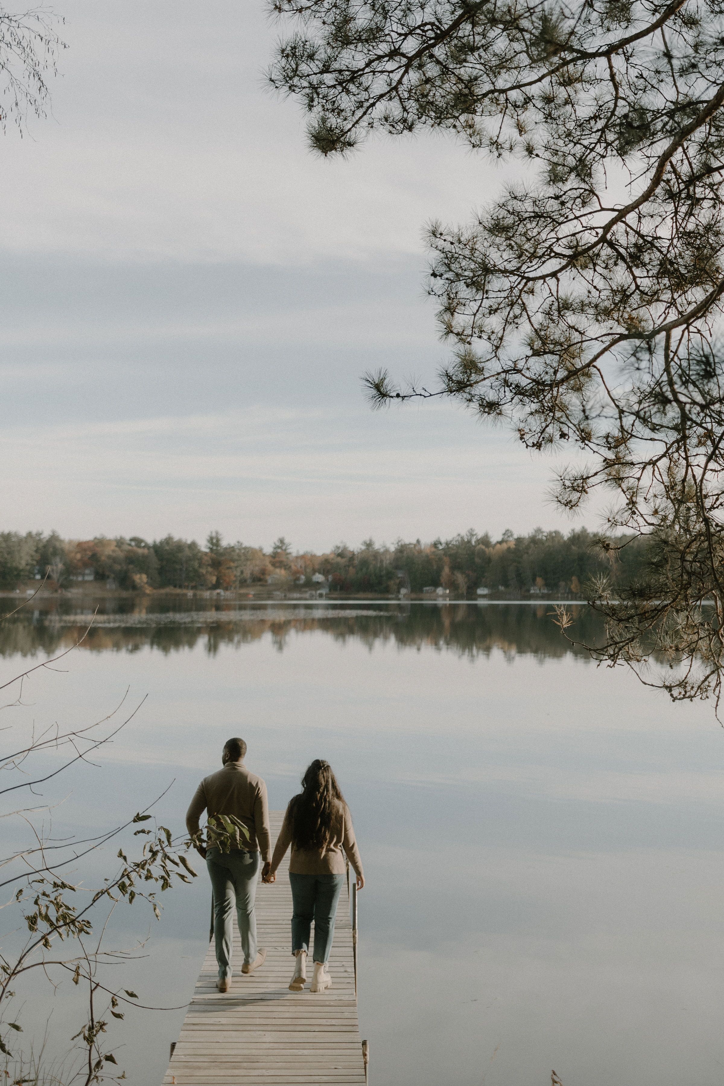 Una pareja caminando sobre un muelle de madera sobre un lago tranquilo, rodeada de árboles.